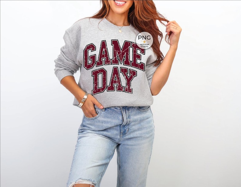 May include: A heather gray sweatshirt with the words "GAME DAY" in a distressed maroon font. The sweatshirt has long sleeves and a crew neck. The text is in a bold, collegiate style. The sweatshirt is paired with blue jeans.