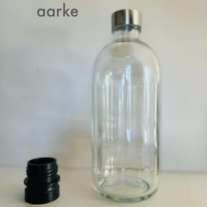 Könnte beinhalten: Eine klare Glasflasche mit einem silbernen Deckel und einem schwarzen Gummistopfen. Die Flasche hat eine strukturierte Oberfläche und das Wort "aark" ist auf dem Hintergrund aufgedruckt.