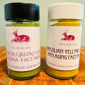 Puede incluir: Dos frascos de vidrio con etiquetas blancas y tapas negras. Las etiquetas tienen un logotipo de ciervo y el texto "VEDA PURE SKIN". El frasco de la izquierda contiene una mascarilla facial de arcilla verde y el texto "French Green Clay Pulina Face Mask" y "With Rosehip, Jojoba Oil". El frasco de la derecha contiene una mascarilla facial de arcilla amarilla y el texto "Brazilian Yellow Clay Anti Aging Face Mask" y "Detoxing - Exfoliating - Moisturizing".