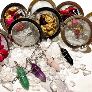 May include: Five stainless steel tea infusers containing various dried flowers and herbs. Four crystal pendants with chains are scattered around the infusers.