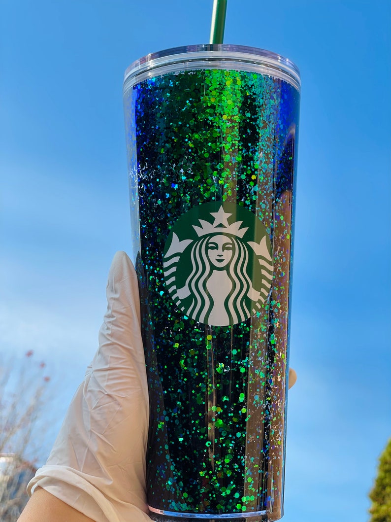 Pu&ograve; includere: Un bicchiere trasparente con un design di glitter verde e blu e un logo Starbucks. Il bicchiere ha una cannuccia.