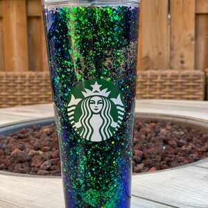 Pu&ograve; includere: Un bicchiere trasparente con un design di glitter verde, blu e nero. Il bicchiere ha il logo Starbucks sul davanti.