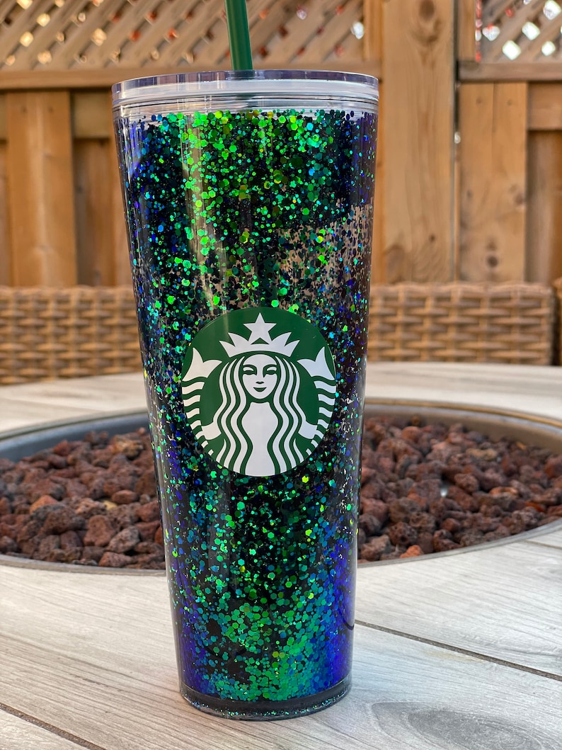 Pu&ograve; includere: Un bicchiere trasparente con un design di glitter verde, blu e nero. Il bicchiere ha il logo Starbucks sul davanti.