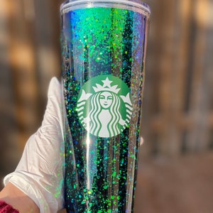 Pu&ograve; includere: Un bicchiere trasparente con un design di glitter nero e verde. Il bicchiere ha un coperchio verde e una cannuccia verde. Il logo Starbucks &egrave; stampato sul davanti del bicchiere.