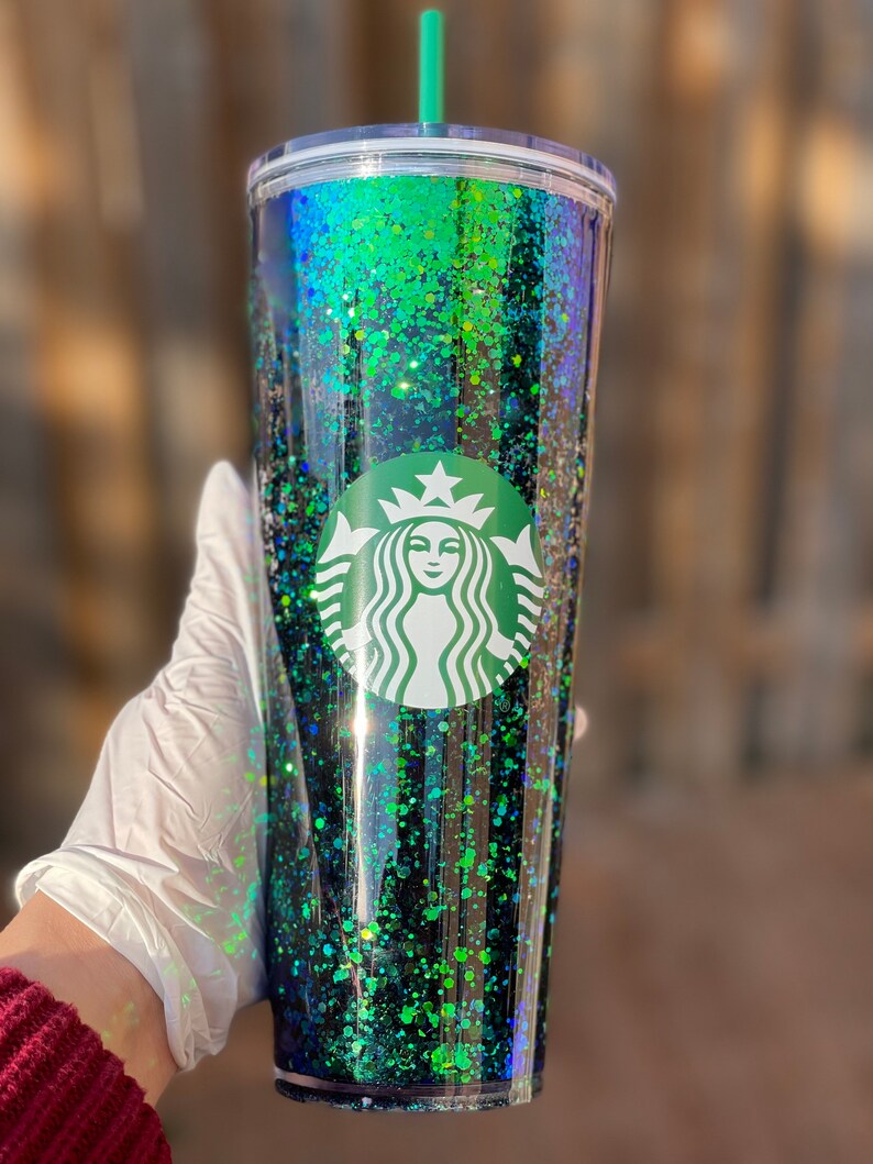 Pu&ograve; includere: Un bicchiere trasparente con un design di glitter nero e verde. Il bicchiere ha un coperchio verde e una cannuccia verde. Il logo Starbucks &egrave; stampato sul davanti del bicchiere.