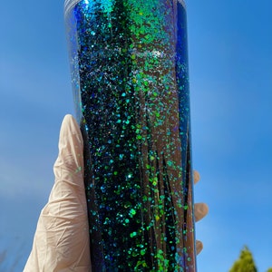 Pu&ograve; includere: Un bicchiere di plastica trasparente con un design di glitter verde e blu. Il bicchiere ha un coperchio e una cannuccia.
