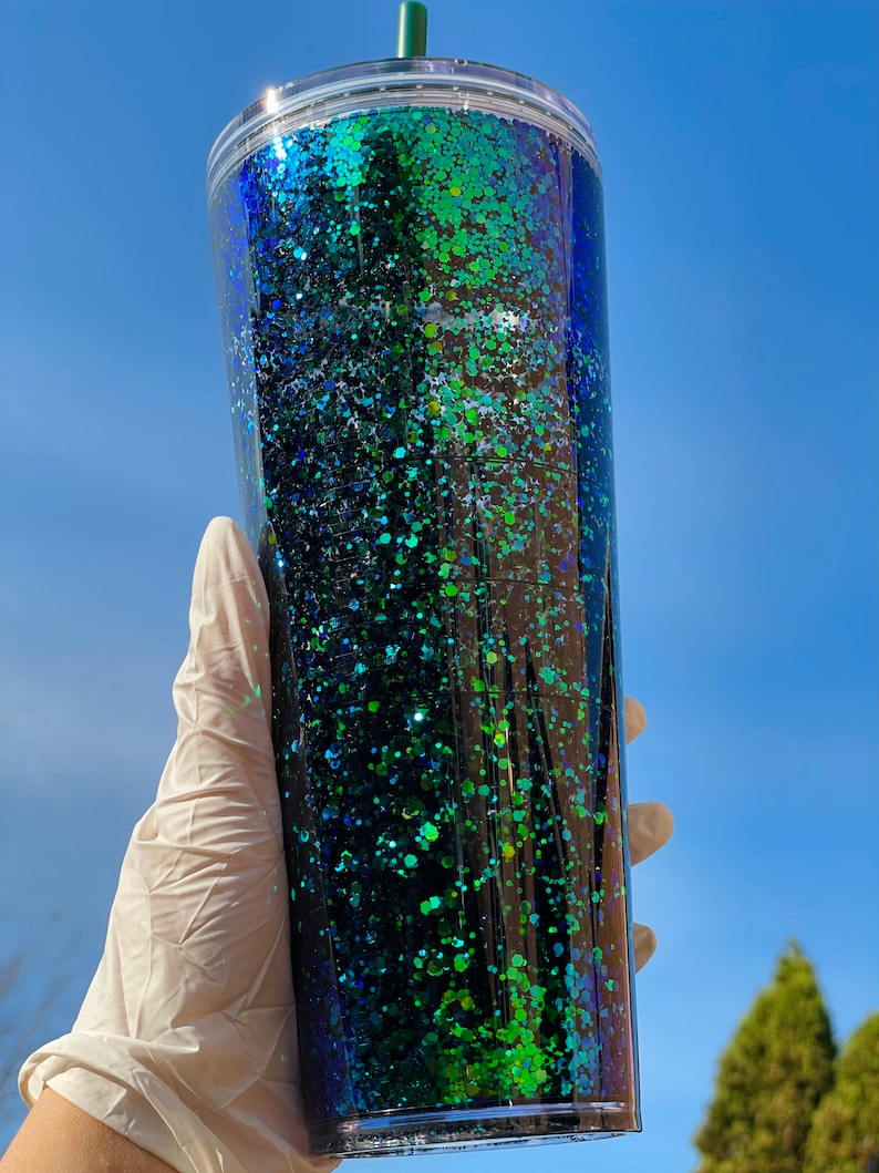 Pu&ograve; includere: Un bicchiere di plastica trasparente con un design di glitter verde e blu. Il bicchiere ha un coperchio e una cannuccia.