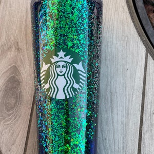 Pu&ograve; includere: Un bicchiere trasparente con un design di glitter verde e nero. Il bicchiere ha il logo Starbucks sul davanti.