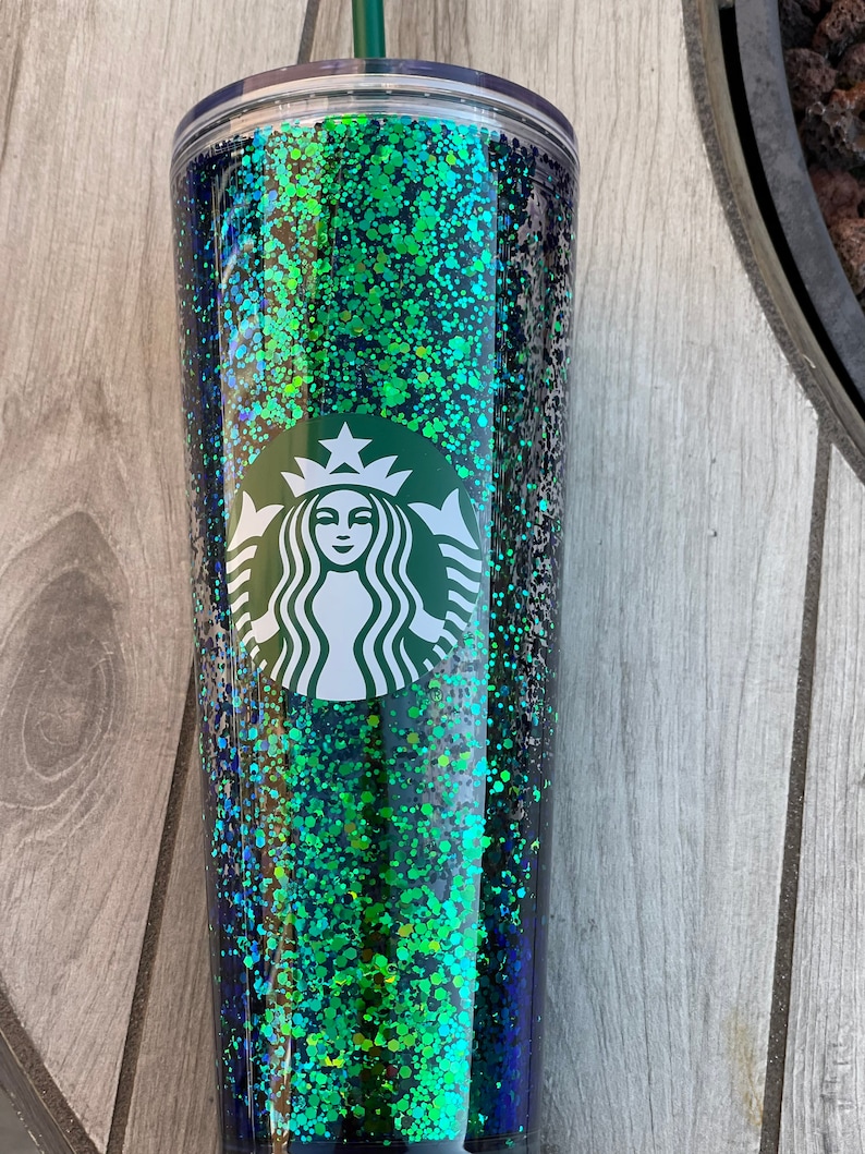 Pu&ograve; includere: Un bicchiere trasparente con un design di glitter verde e nero. Il bicchiere ha il logo Starbucks sul davanti.