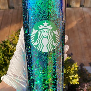 Pu&ograve; includere: Un bicchiere di glitter nero e verde con il logo Starbucks. Il bicchiere ha un coperchio trasparente e una cannuccia.