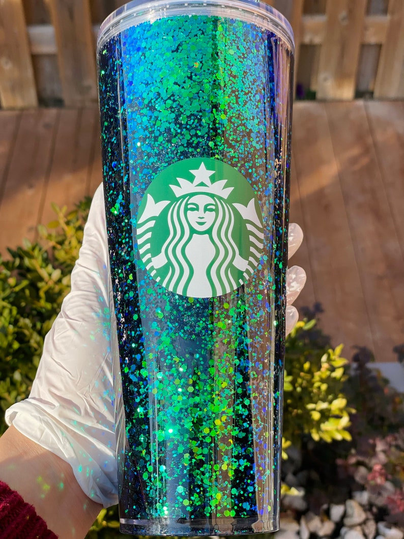 Pu&ograve; includere: Un bicchiere di glitter nero e verde con il logo Starbucks. Il bicchiere ha un coperchio trasparente e una cannuccia.