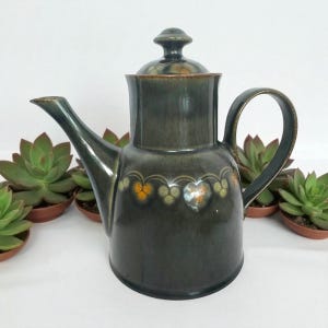 May include: Dark green ceramic coffee pot with a lid and handle. The pot has a decorative band of yellow and orange floral designs around the middle. The spout is long and curved. The pot is surrounded by small potted succulents.