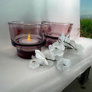 May include: Two purple glass candle holders with a white tea light candle in one of the holders. The candle holders are on a white surface with white flowers.