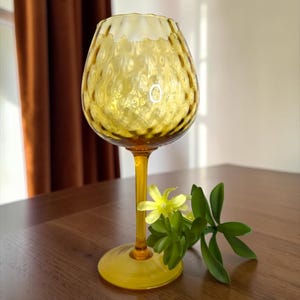May include: A yellow glass goblet with a textured, bubble-like pattern on the bowl. The goblet has a long stem and a round base. A small sprig of green leaves and yellow flowers rests beside the goblet on a wooden surface.
