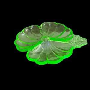 May include: A translucent, light green glass dish shaped like a flower with scalloped edges and a leaf-shaped handle. The dish features a segmented interior with raised ridges radiating from the center, creating a distinct floral pattern.