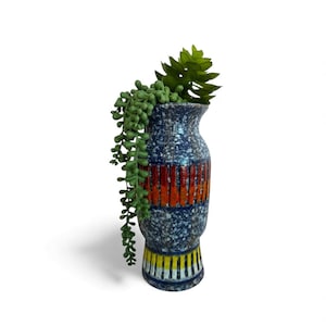 May include: A tall, ceramic vase with a speckled blue background and vertical orange stripes. The base features yellow and white stripes. The vase holds green, leafy plants, creating a decorative display.