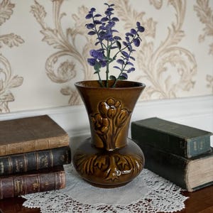 May include: A brown ceramic vase with a bulbous base and flared top, featuring embossed floral designs. The vase holds purple artificial flowers. It sits on a white lace doily, with antique books in the background.