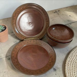 May include: A set of brown ceramic plates and bowls. Two large plates and two small bowls are arranged on a wooden surface. The plates have a decorative border. A small terracotta pot with a succulent plant is on the left.