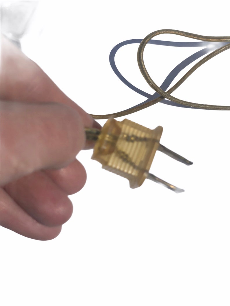 Puede incluir: Primer plano de un enchufe el&eacute;ctrico dorado con dos clavijas. El enchufe est&aacute; conectado a un cable el&eacute;ctrico marr&oacute;n.