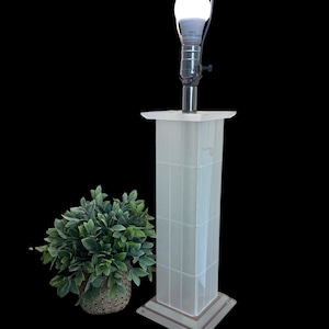 May include: A white, rectangular table lamp with a grid pattern on the column. The lamp has a silver neck and a bright white LED light bulb. A small potted plant with green leaves sits to the left of the lamp.