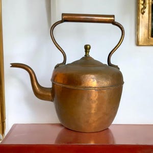 May include: A vintage copper kettle with a brass handle and spout. The kettle has a rounded body and a polished finish.