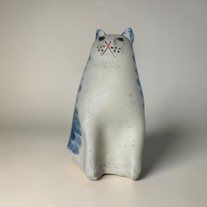 May include: A ceramic cat figurine with a speckled off-white body and blue painted stripes along the sides. The cat has a pointed face with small dark eyes, a pink nose, and a series of small dots representing whiskers on its muzzle.