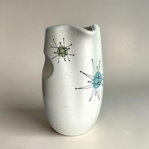 May include: A white ceramic pitcher with a speckled finish, featuring black starburst line patterns overlaid on light green and light blue circular accents. The vessel has an asymmetrical, curved rim and a smooth, rounded body shape.