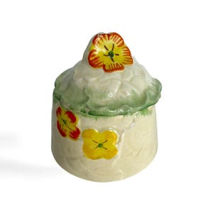 May include: A small ceramic jar with a lid decorated with yellow, orange, and green flowers. The jar has a white background with a textured surface.