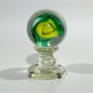 May include: A clear glass paperweight with a spherical top featuring a swirl of green and yellow colors. The paperweight sits on a clear, pedestal-style base with a circular foot. The base has small, silver-colored accents.
