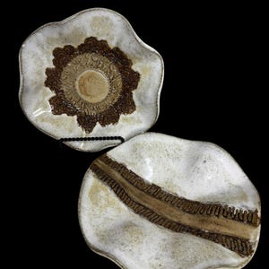 May include: Two ceramic dishes with wavy, irregular edges and a cream-colored glaze. One dish features a central brown circular pattern with intricate detailing, while the other displays a wide, textured brown band running across its center.