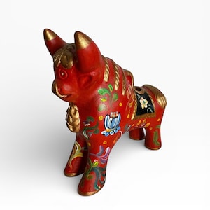 May include: A red ceramic bull figurine with gold accents on the horns, mane, and hooves. The bull is decorated with colorful floral patterns in blue, green, and yellow. The figurine has a black saddle with floral details.
