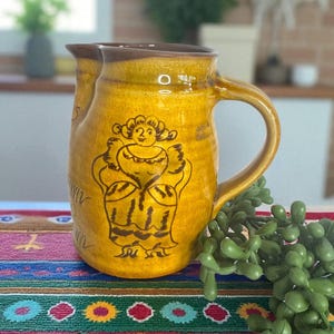 May include: A handmade ceramic pitcher with a yellow glaze and a brown rim and handle. The pitcher features a hand-painted illustration of a woman in a dress. The pitcher is sitting on a colorful patterned textile.