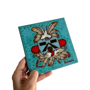 May include: Square ceramic tile with a turquoise background and a Native American-inspired design. The design features a stylized face with black, red, and white accents, and feather details. The tile is held in a hand.