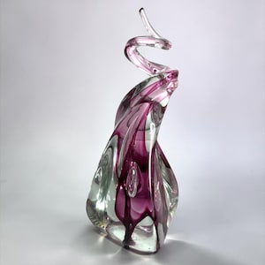 May include: A decorative glass sculpture with a swirling, abstract design. The sculpture features a clear base with streaks of deep pink and a clear, spiraling top. The glass is shaped into flowing curves, creating a dynamic and artistic form.