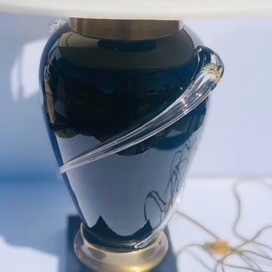 Puede incluir: Una l&aacute;mpara de mesa de vidrio negro con una pantalla blanca y un dise&ntilde;o de vidrio transparente. La l&aacute;mpara tiene una base dorada y una base cuadrada negra.