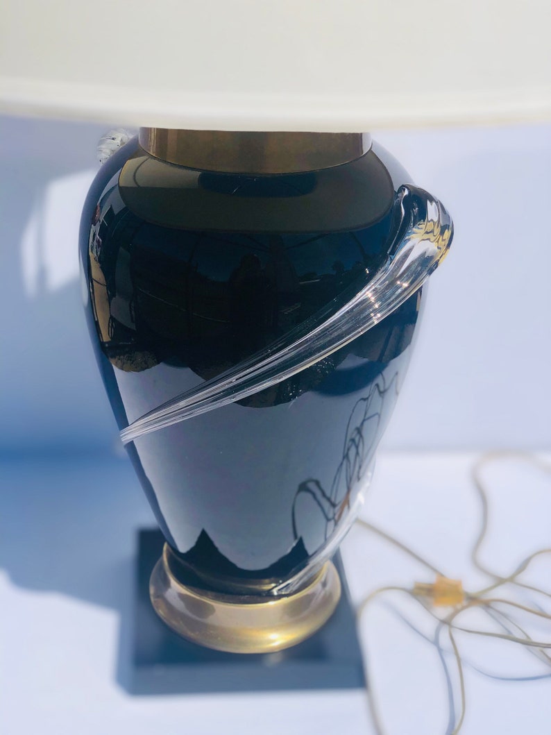 Puede incluir: Una l&aacute;mpara de mesa de vidrio negro con una pantalla blanca y un dise&ntilde;o de vidrio transparente. La l&aacute;mpara tiene una base dorada y una base cuadrada negra.