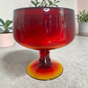 May include: A vintage red glass goblet with a wide bowl and a short stem. The base of the goblet is a gradient of yellow to orange. The glass is transparent, allowing light to pass through. The goblet is likely used for serving beverages.