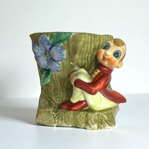 May include: A vintage ceramic planter shaped like a tree trunk, featuring a seated elf figure. The elf wears a red jacket, yellow pants, and red shoes. A blue flower with green leaves decorates the side of the planter. A whimsical decorative item.