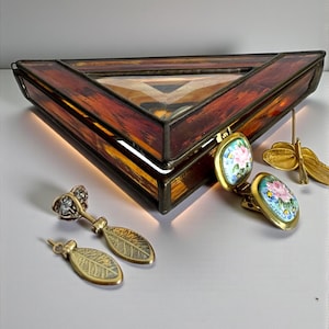 May include: A triangular stained glass jewelry box with a warm amber and brown color scheme.  Accompanying the box are several pieces of jewelry, including gold-toned leaf earrings, floral clip-on earrings, and a dragonfly brooch.