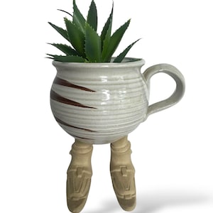 Puede incluir: Una maceta de cerámica caprichosa con forma de taza, con asa y patas. La taza blanquecina presenta rayas marrones y contiene una planta suculenta verde. Las patas son de color beige, lo que añade un toque único a esta pieza decorativa.