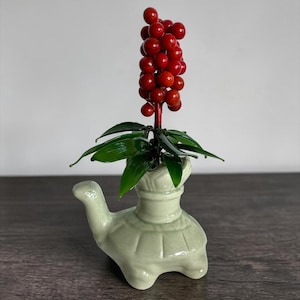 May include: A light green ceramic turtle-shaped vase containing a decorative plant with dark green leaves and a cluster of bright red berries. The turtle figure has a smooth, glazed finish and a distinct shell pattern on its back.