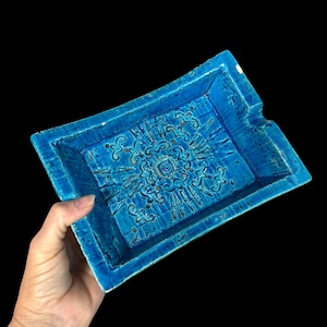 May include: A rectangular turquoise ceramic tray featuring a central embossed floral and geometric pattern with a deep blue glaze. The tray has raised edges and a notched corner, showcasing a textured, crackled finish across the surface.