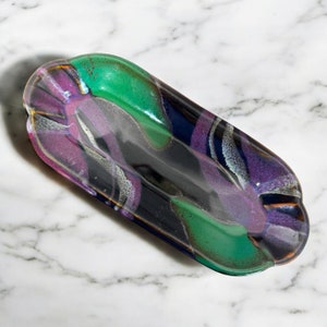 May include: An elongated oval glass tray featuring a glossy finish with a swirling abstract pattern of deep black, vibrant green, and rich purple hues. The surface displays a smooth, reflective texture with blended color transitions throughout.