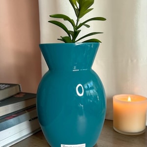 May include: A teal-colored Haeger Pottery vase with a glossy finish, holding a green plant. The vase has a rounded body and a flared top. A lit candle and books are in the background.