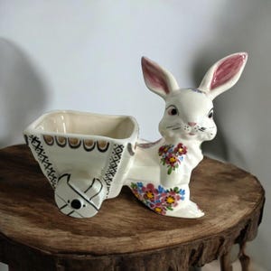 May include: A ceramic bunny planter with a cart, painted in cream, brown, and floral accents. The bunny has pink-lined ears and is decorated with colorful flowers. The cart has a square opening for plants.