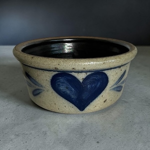 May include: A small, round, stoneware bowl with a dark interior and a light beige exterior. The bowl features a large, blue heart design on the front, with blue leaf accents on either side. The rim is a darker beige color.