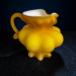 May include: A small, vibrant yellow glass creamer pitcher with a satin, frosted finish. The vessel features a bulbous, rounded body with a ruffled, scalloped rim and a curved handle. The interior of the pitcher is a smooth, opaque white glass.