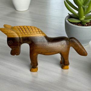 May include: A handcrafted wooden horse figurine. The horse is carved from wood with a natural grain, showcasing shades of brown and tan. The mane features carved lines, and the horse stands on four small legs. A decorative item.