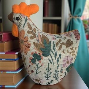 May include: A decorative chicken-shaped doorstop. The doorstop features a floral pattern in shades of orange, green, and pink on a cream background. It has an orange comb and wattle. The doorstop is made of fabric and sits on a wooden surface.