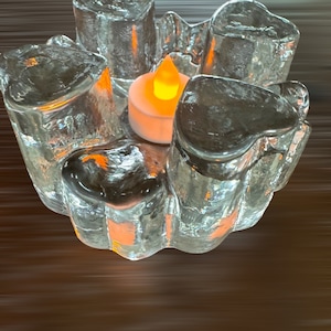 May include: A clear glass candle holder with a textured surface. The holder has five sections, each shaped like a heart. A small white candle is burning inside the holder.